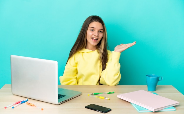 Klein meisje in een tafel met een laptop over een geïsoleerde blauwe achtergrond met een denkbeeldige kopie op de handpalm om een advertentie in te voegen