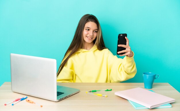 Klein meisje in een tafel met een laptop over geïsoleerde blauwe achtergrond die een selfie maakt