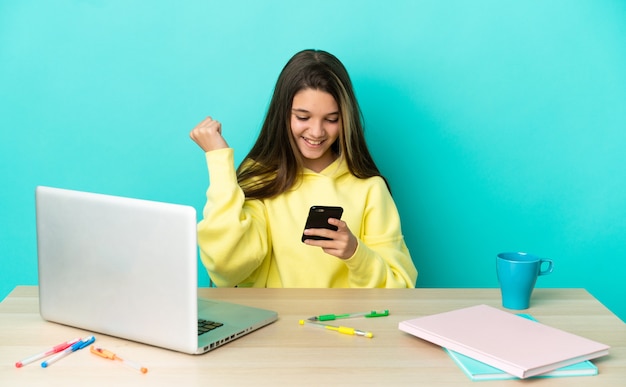 Klein meisje in een tafel met een laptop over geïsoleerde blauwe achtergrond verrast en stuurt een bericht