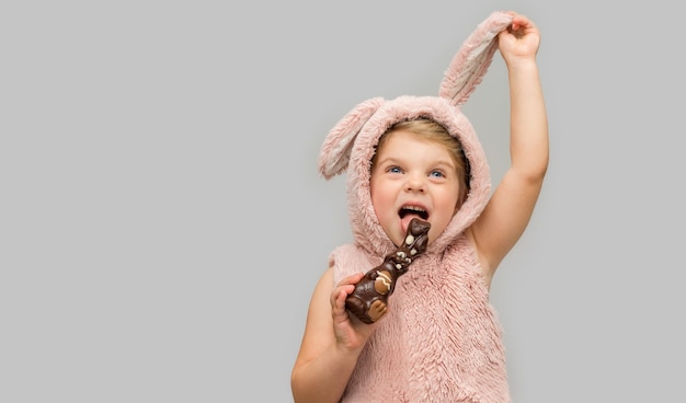 Klein meisje in een vest met konijnenoren eet heerlijk een chocoladehaas op Paasavond Grijze achtergrond met plaats voor tekst Verkoopkortingen winkelen voor de vakantie