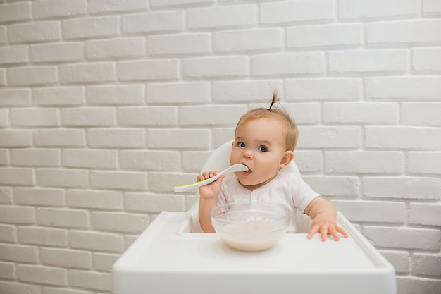 klein meisje in een witte bodysuit zit in een voederstoel met een lepel