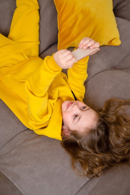 klein meisje in gele kleren ligt op haar rug op het bed en houdt een smartphone in haar handen