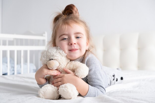 Klein meisje in grijze coltrui met teddy beer witte beddengoed glimlachen opleggen.