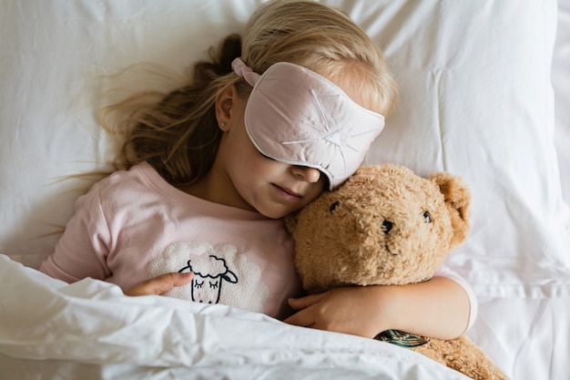 klein meisje in pyjama en blinddoek slapen in witte bed met teddybeer