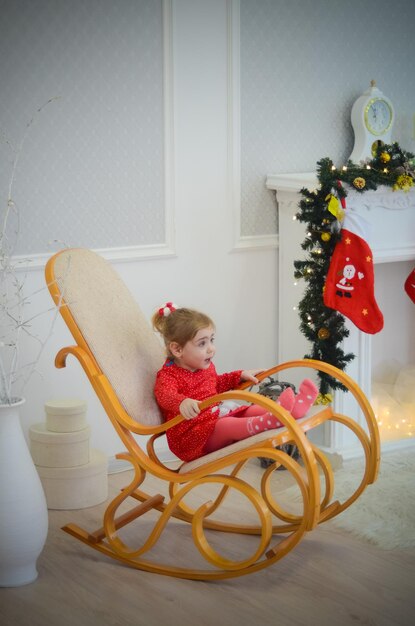 Klein meisje in rode jurk spelen in de kamer met kerstversieringen in een schommelstoel