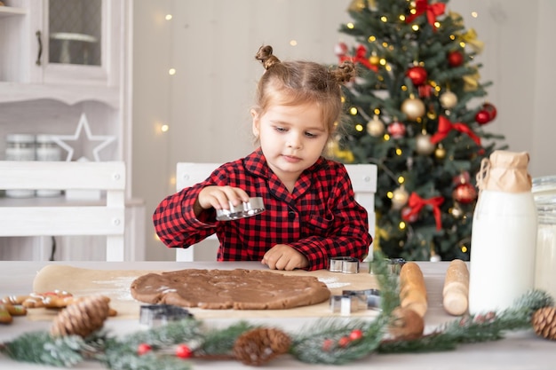 Klein meisje in rode pyjama die feestelijke peperkoek kookt in met kerst versierde keuken