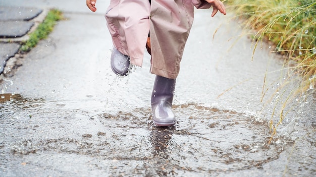 Klein meisje in roze waterdichte regenjas paarse rubberen laarzen grappige springt door plassen op straat weg in regenachtige dag weer Lente herfst Kinderplezier na regen Outdoor recreatie activiteit