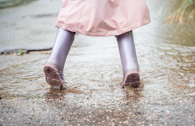 Klein meisje in roze waterdichte regenjas paarse rubberen laarzen grappige springt door plassen op straat weg in regenachtige dag weer lente herfst kinderplezier na regen outdoor recreatie activiteit