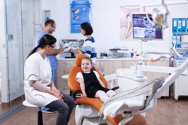 Klein meisje in stomatologiebureau voor tandenbehandeling die tandarts bekijkt. Kind met haar moeder tijdens tandencontrole met stomatolog zittend op een stoel.