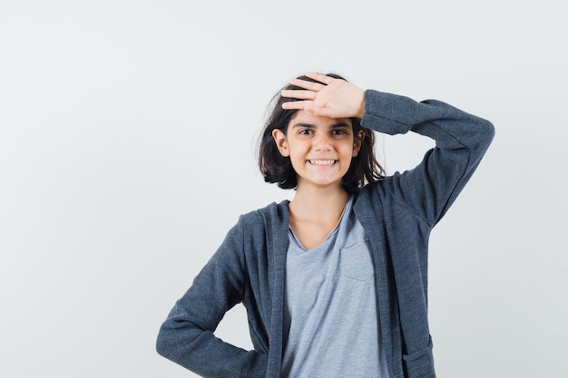 Klein meisje in t-shirt, jasje met hand op het voorhoofd en op zoek vrolijk, vooraanzicht.