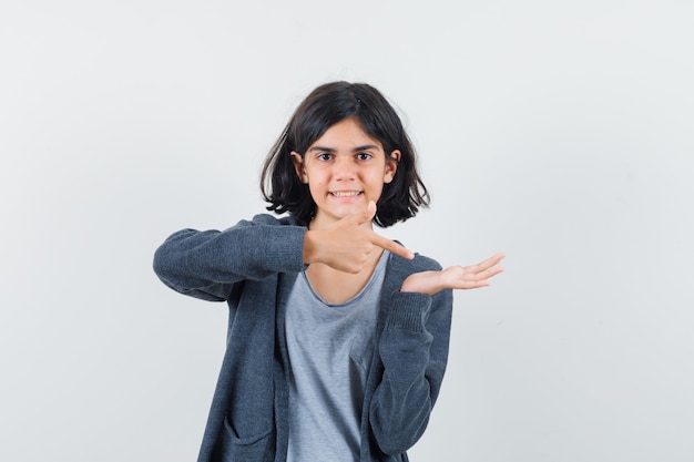 Klein meisje in t-shirt, jasje wijzend op haar lege handpalm en kijkt zelfverzekerd, vooraanzicht.