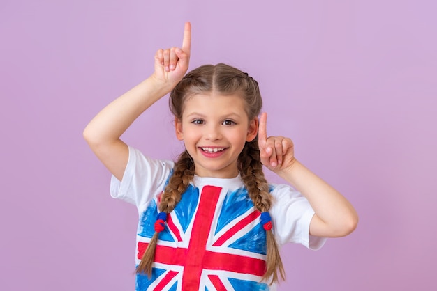 Klein meisje in t-shirt met vlag van Groot-Brittannië wijst omhoog op lichtpaarse achtergrond