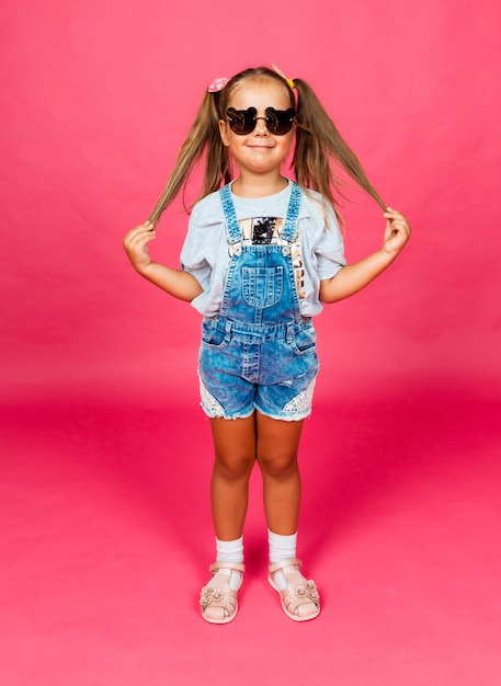 Klein meisje in zonnebril kijken naar de camera met een glimlach, staande op een roze achtergrond. kinderen.