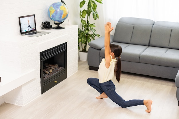 Klein meisje kijkt naar online video op laptop en doet thuis workout- of fitnessoefeningen in haar kamer. Online training op afstand, zelfisolatie op sociale afstand, online onderwijsconcept