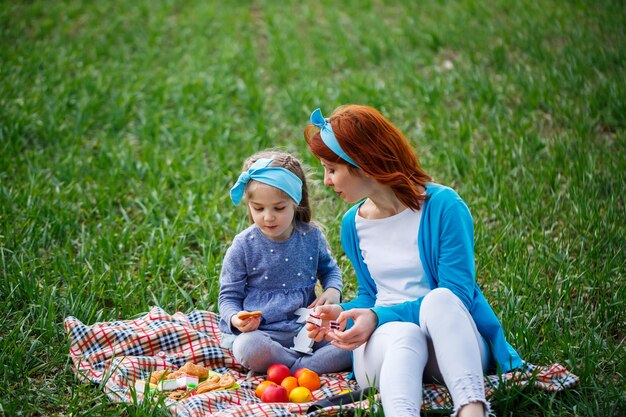 Klein meisje kind en moeder vrouw zittend op de sprei en koekjes en marmelade eten, groen gras in het veld, zonnig lenteweer, glimlach en vreugde van het kind, blauwe lucht met wolken