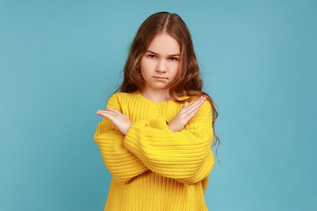 Klein meisje kruist handen kijkend naar camera met agressie tonen stop gebaar manier verboden dragen gele casual stijl trui Indoor studio shot geïsoleerd op blauwe achtergrond