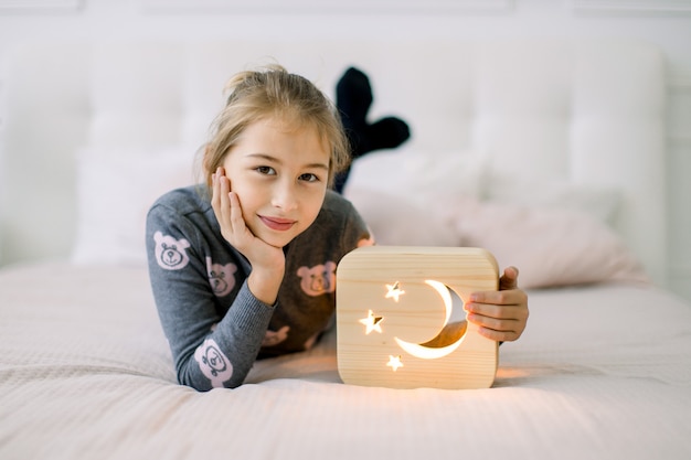 klein meisje liggend op het bed op gezellige lichte slaapkamer thuis met houten stijlvolle nachtlamp met uitgesneden maan en sterren foto