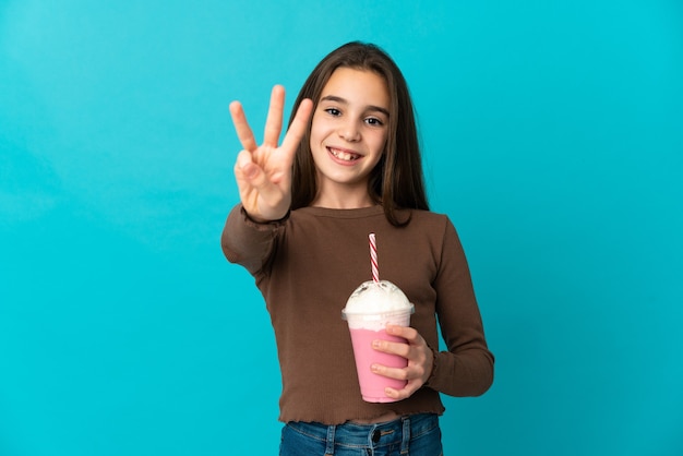 Klein meisje met aardbeienmilkshake geïsoleerd op blauwe achtergrond gelukkig en drie tellen met vingers