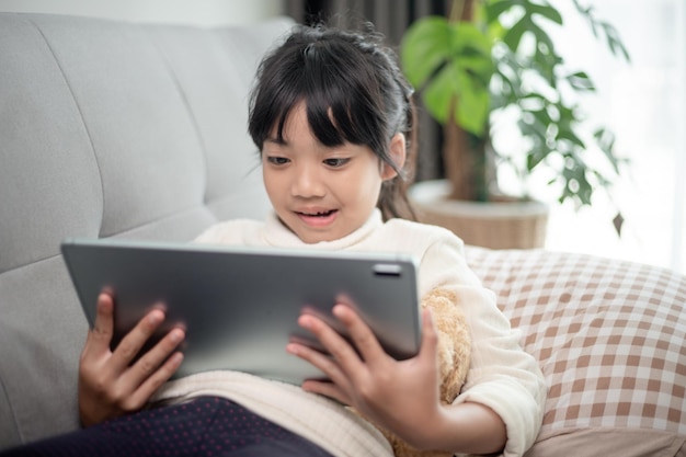 Klein meisje met behulp van tablet speelspel op internet Kind zittend op de bank kijken of praten met een vriend online Kind ontspannen in de woonkamer in de ochtend Kinderen met nieuwe technologie concept