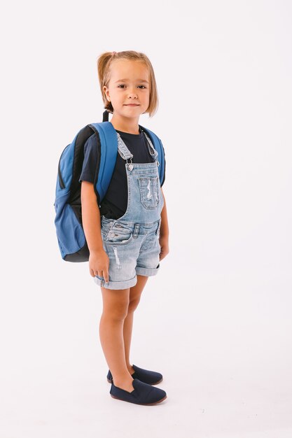 Klein meisje met blond haar gekleed in een blauwe tuinbroek en t-shirt, met een rugzak klaar voor terug naar school, op witte achtergrond