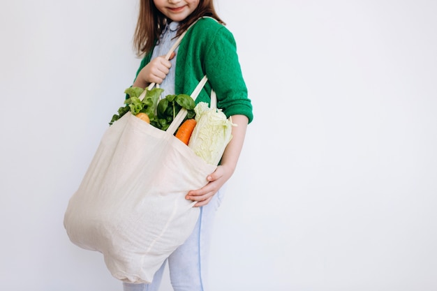 Klein meisje met een boodschappentas van textiel met groenten. Geen afvalconcept. Pakketvrij boodschappen doen. Eco vriendelijke natuurlijke tas met biologische groenten en fruit.