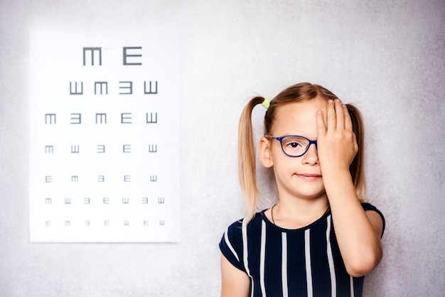 Klein meisje met een bril die haar oog bedekt met één hand die een gezichtstest doet voor school met een wazige ooggrafiek op de achtergrond