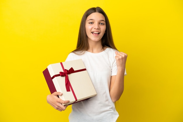 Klein meisje met een geschenk over een geïsoleerde gele achtergrond die een overwinning viert in winnaarspositie