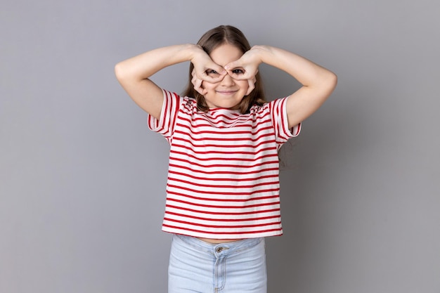 Klein meisje met een gestreept t-shirt dat een verrekijkergebaar maakt en ver weg kijkt met een schattige glimlach