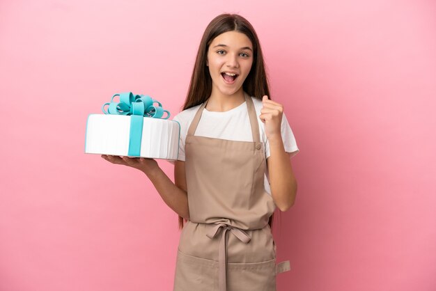 Klein meisje met een grote taart over geïsoleerde roze muur die een overwinning viert in winnaarspositie