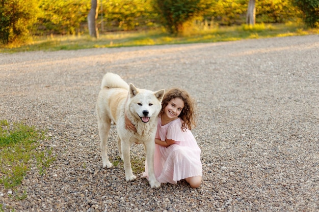 Klein meisje met een grote witte hond in het park Een mooi 7-jarig meisje in roze jurk knuffelt haar favoriete hond tijdens een zomerse wandeling