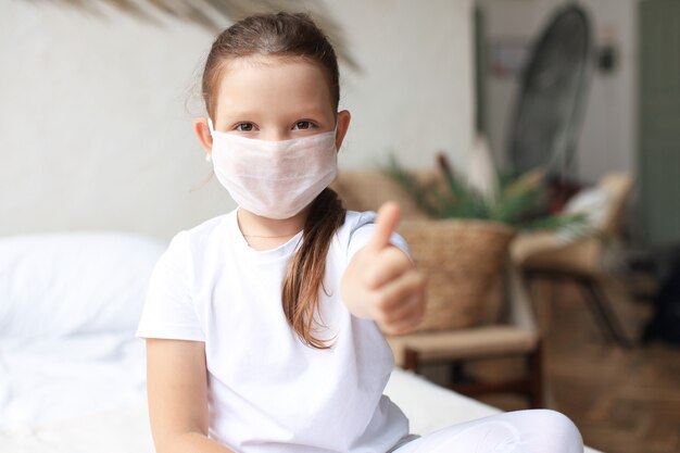 Klein meisje met een medisch masker dat een duim omhoog gebaar toont. Bescherming tegen het coronavirus.