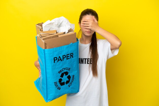 Klein meisje met een recyclingzak vol papier om te recyclen over een geïsoleerde gele achtergrond die de ogen bedekt met de handen. Wil je iets niet zien