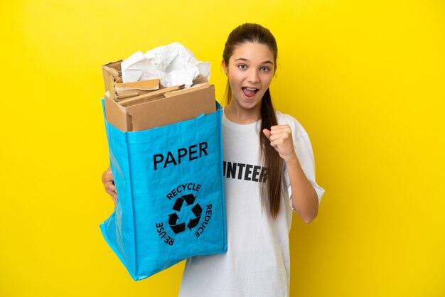 Klein meisje met een recyclingzak vol papier om te recyclen over geïsoleerde gele achtergrond en viert een overwinning in winnaarspositie