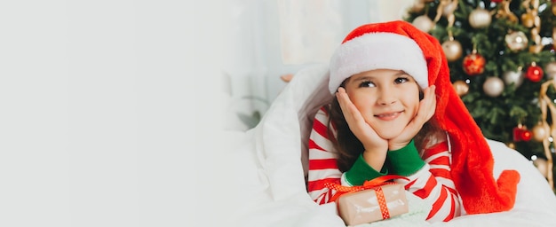 Klein meisje met een rode nieuwjaarshoed met een kerstcadeau Ze ligt op het bed en knuffelt een doos tegen de achtergrond van een kerstboom