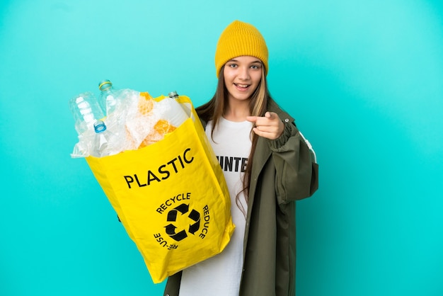 Foto klein meisje met een zak vol plastic flessen om te recyclen over een geïsoleerde blauwe achtergrond, verrast en naar voren wijzend