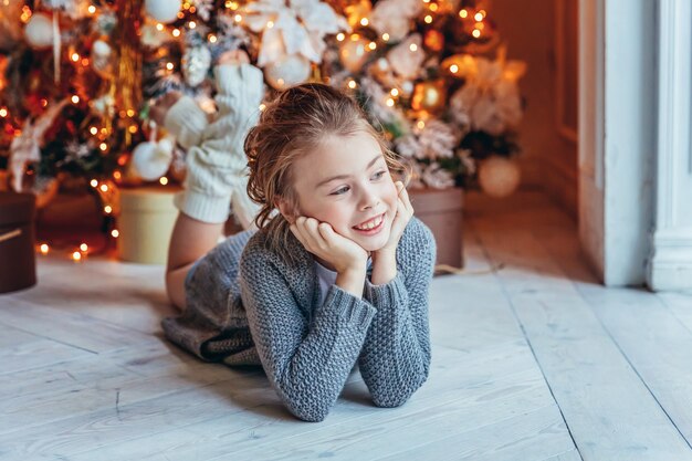 Klein meisje met geschenkdoos in de buurt van kerstboom op kerstavond thuis. Jonge jongen in lichte slaapkamer met winterdecoratie Gelukkig gezin thuis. Kerstmis Nieuwjaar december tijd voor viering concept