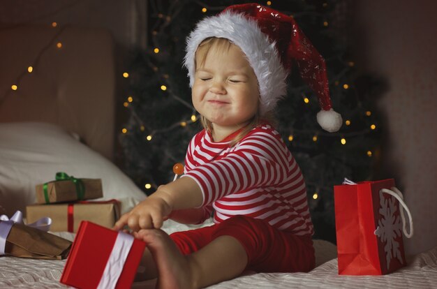 Klein meisje met kerstmuts die de doos van de kerstcadeau opent Grappige en expressieve peuter die van cadeaus geniet