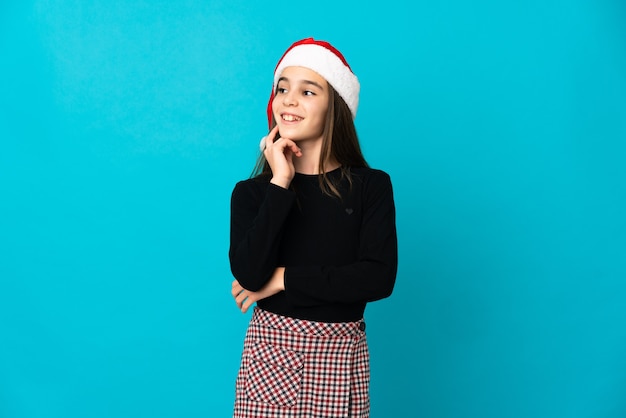 Klein meisje met kerstmuts geïsoleerd op blauwe achtergrond een idee denken tijdens het opzoeken