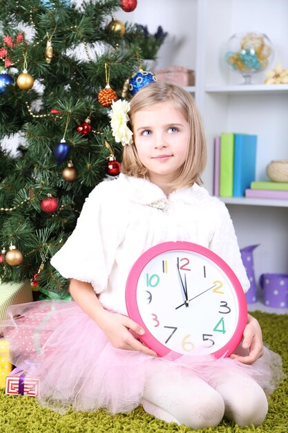 Klein meisje met klok in de buurt van kerstboom in kamer