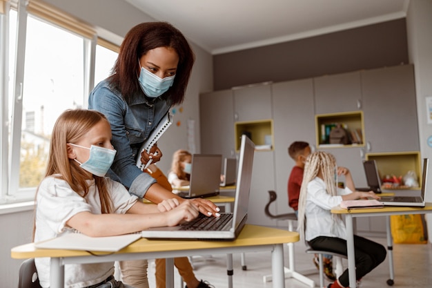 Klein meisje met masker dat laptop gebruikt en aan het bureau in de klas zit