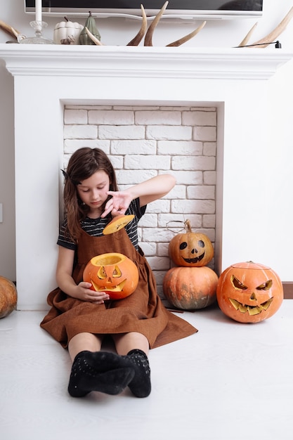 Klein meisje met snijwerk pompoen op Halloween thuis naast de open haard in de woonkamer. Snoep of je leven. Kind dat Halloween viert.