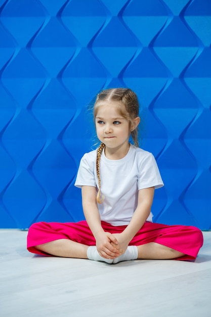 Klein meisje met staartjes in een wit T-shirt op een blauwe achtergrond. Ze zit in lotushouding op de grond en lacht en zwaait met haar handen.