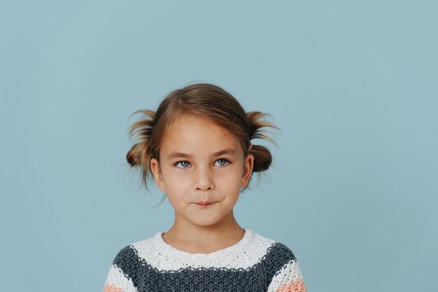 Klein meisje met strakke lippen in gestreepte trui, haar in broodjes op blauwe achtergrond.