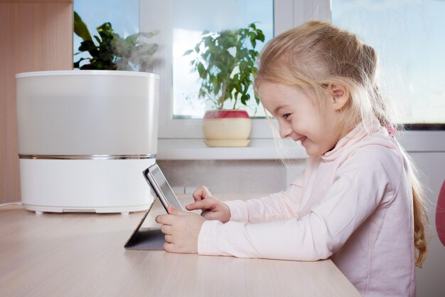 Klein meisje met tablet-pc zittend aan de tafel in de buurt van luchtbevochtiger