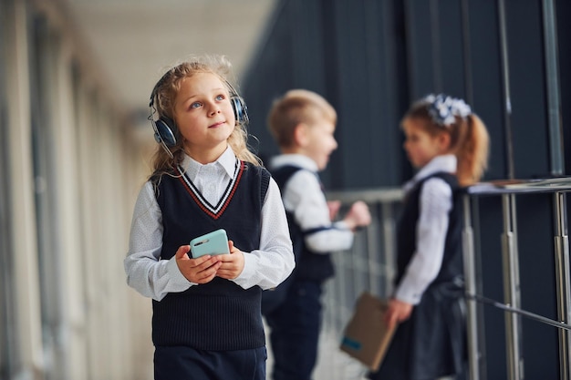 Klein meisje met telefoon en koptelefoon staan voor schoolkinderen in uniform die samen in gang staan Conceptie van onderwijs