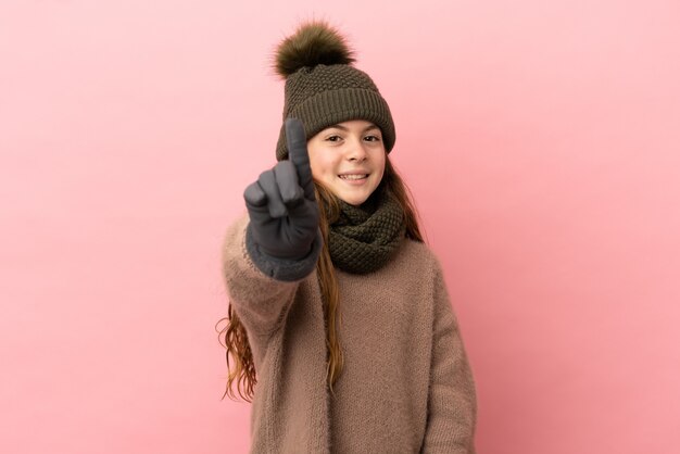 Klein meisje met wintermuts geïsoleerd op roze achtergrond tonen en optillen van een vinger