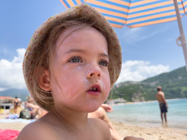 Klein meisje met zonnebrandcrème op haar gezicht zit op het strand onder parasol