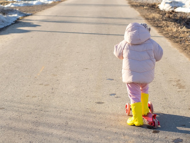 Klein meisje op een scooter in rubberen laarzen in de lente