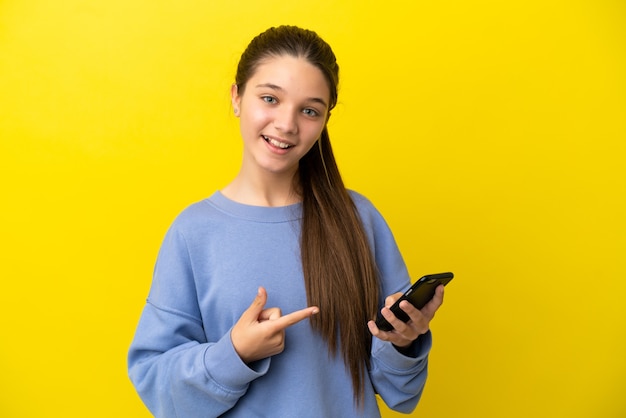 Klein meisje over geïsoleerde gele achtergrond die mobiele telefoon gebruikt en erop wijst