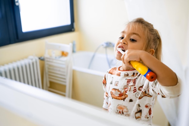klein meisje poetst haar tanden met een elektrische borstel die in de badkamer staat en in de spiegel kijkt